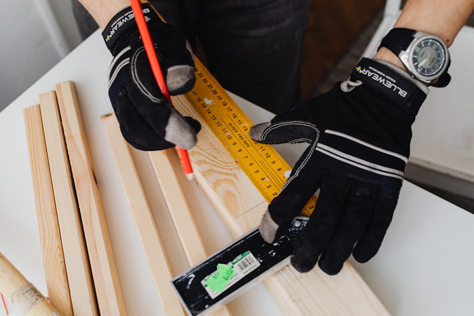 Close-up of a carpenter wearing gloves while measuring a wood plank with a ruler and pencil.